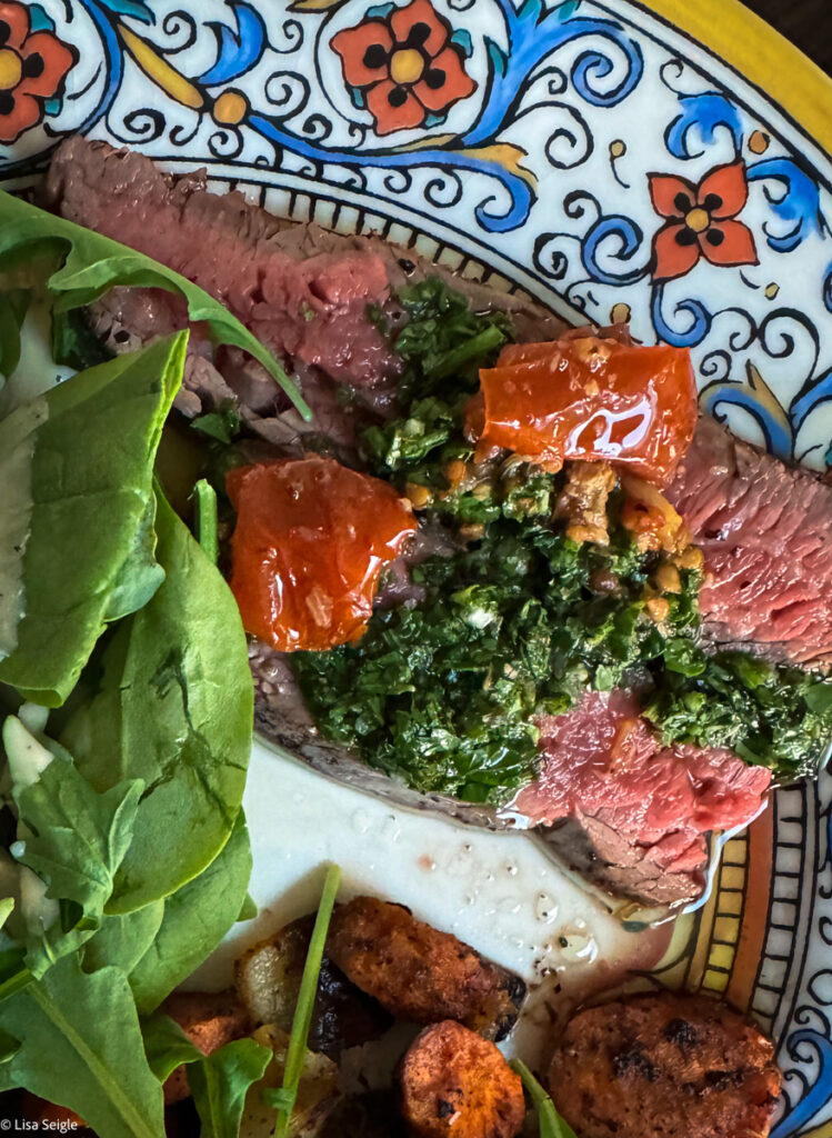 overhead photo of sliced steak on a patterned plate topped with chimichurri and tomatoes