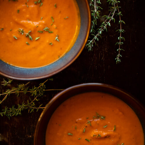 Overhead photo of two bowls of creamy roasted tomato soup on dark background with sprigs of thyme