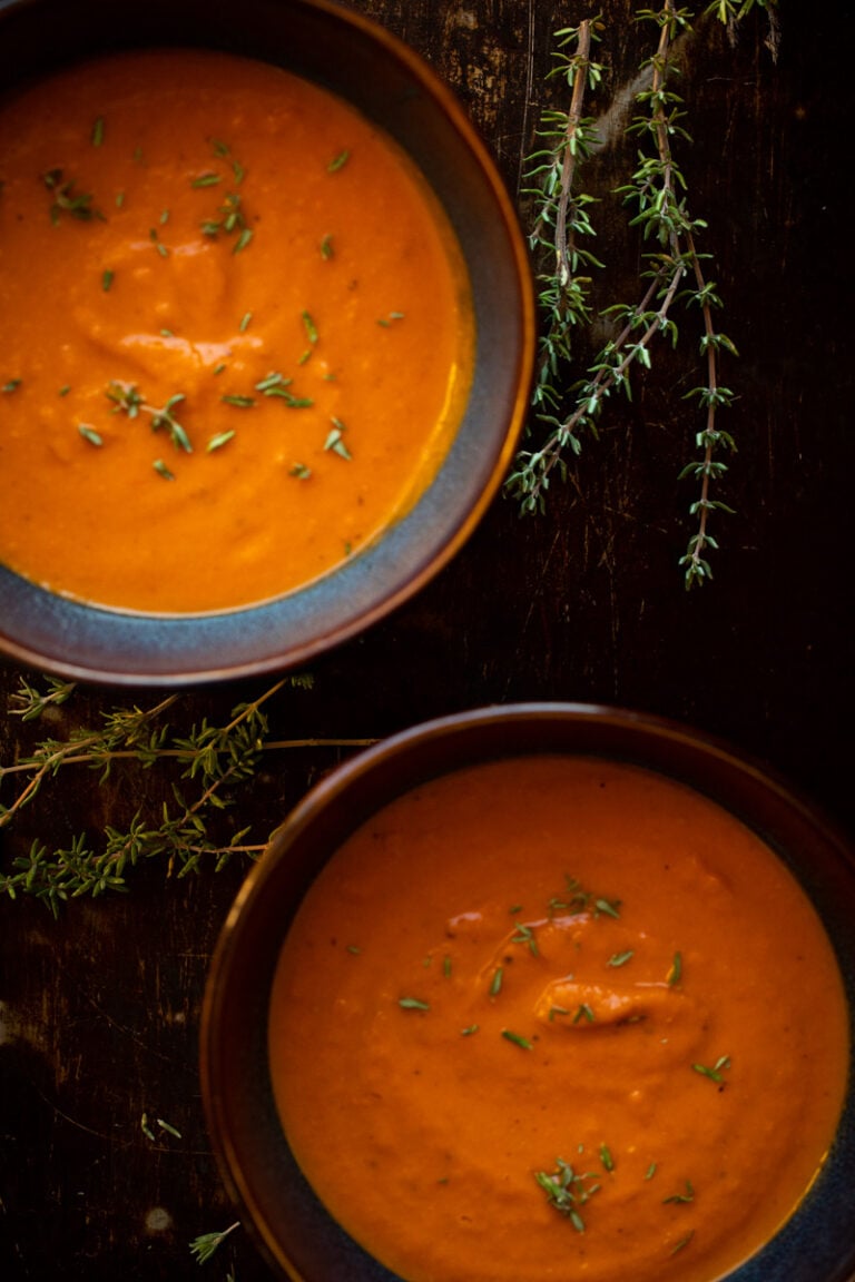 Overhead photo of two bowls of creamy roasted tomato soup on dark background with sprigs of thyme