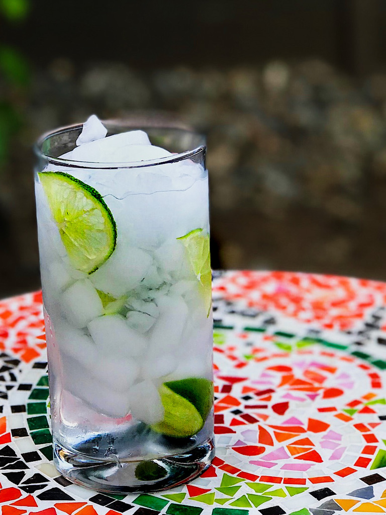 Side view of cucumber gin spritz in a tall glass with slices of cucumber on a colored tile table.