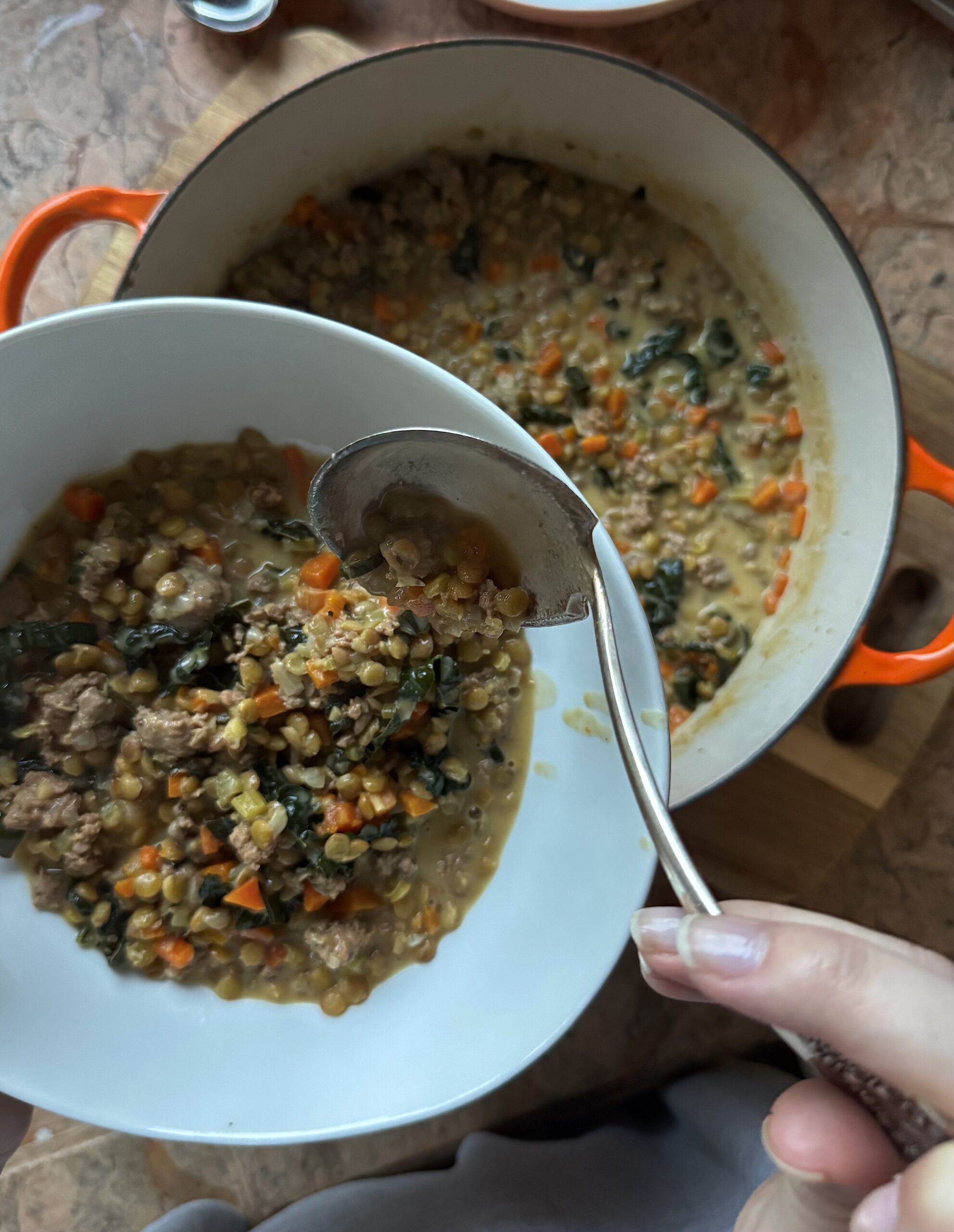Overhead photo of sausage lentil soup being served in a white bowl with a ladle and a large bowl of soup in the background