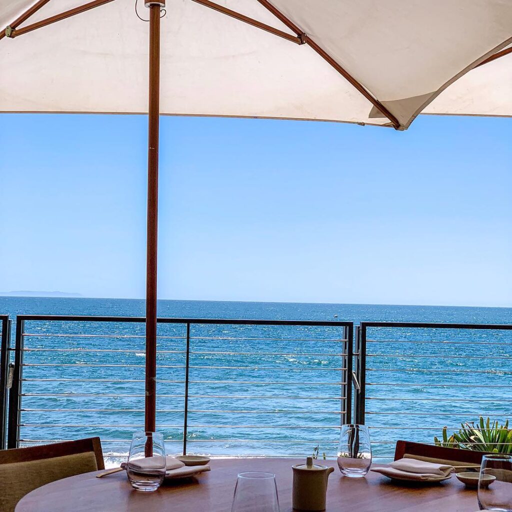 photo of outdoor table set with plates, napkins and glasses with an umbrella on a deck with a view of the ocean