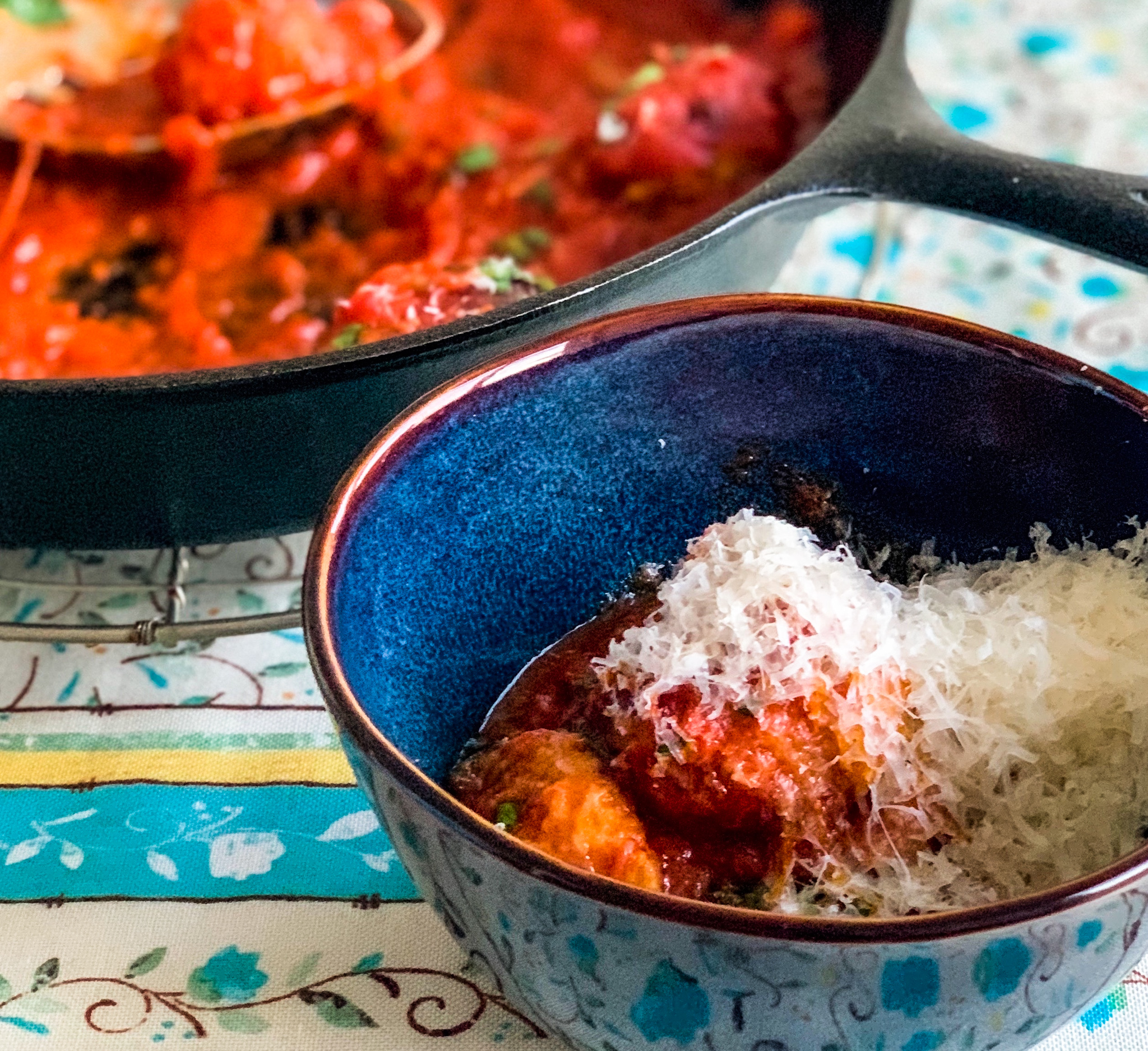 Overhead photo of meatballs in a blue bowl with cheese and a skillet with more meatballs in the background