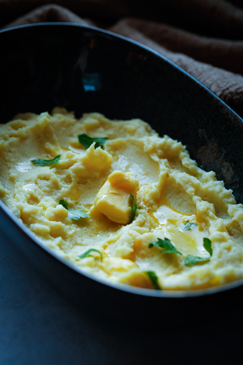 Overhead of mashed potatoes in dark bowl with pad of melted butter and parsley