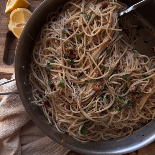 spaghetti with walnuts and anchovies in skillet on counter next to sliced lemon and beige napkin.
