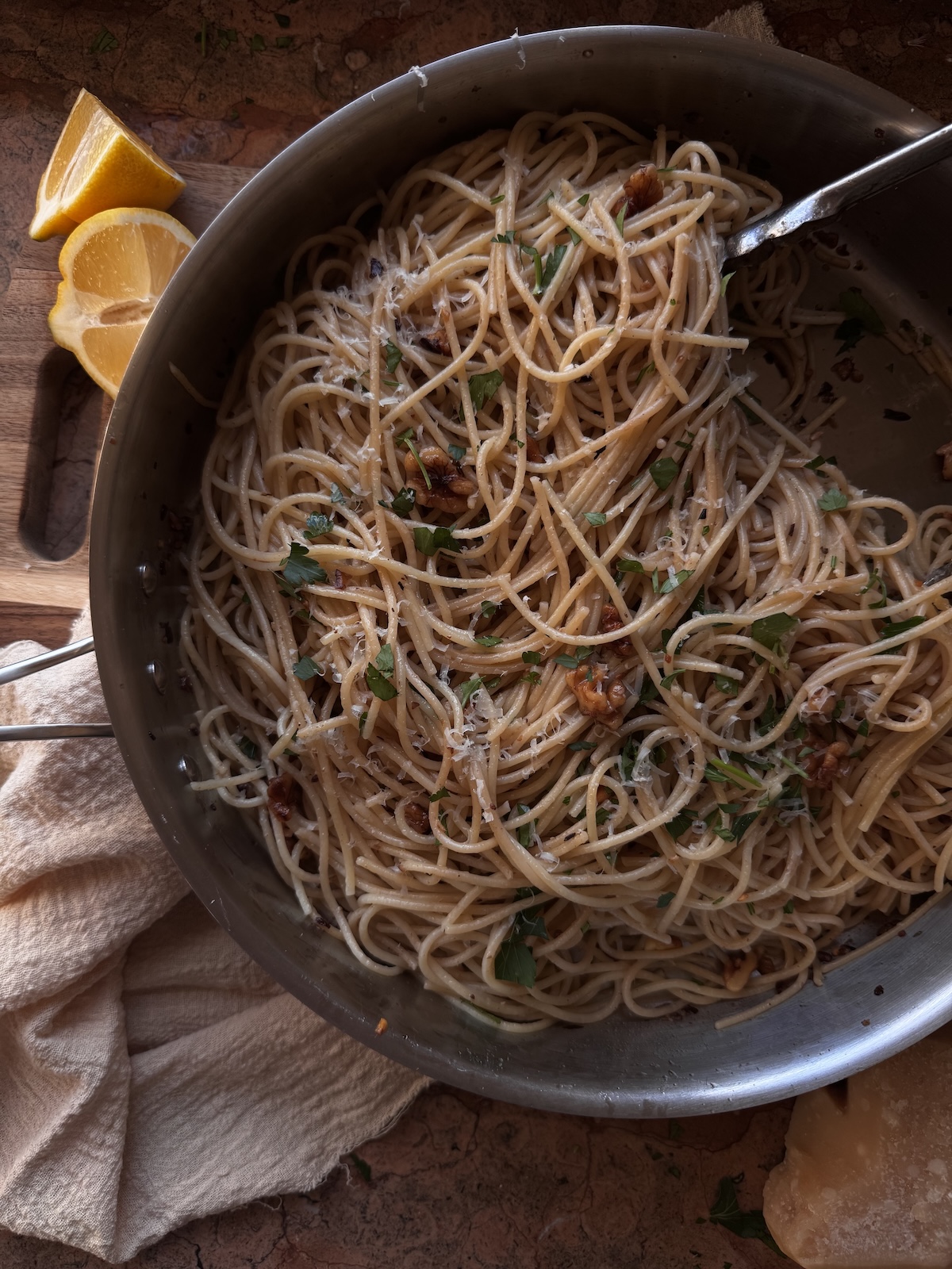 spaghetti with walnuts and anchovies in skillet on counter next to sliced lemon and beige napkin.
