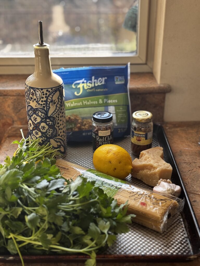 Bottle of oil, walnuts, anchovies, garlic, lemon, parmesan cheese, spaghetti and parsley on cooking sheet in front of a window.
