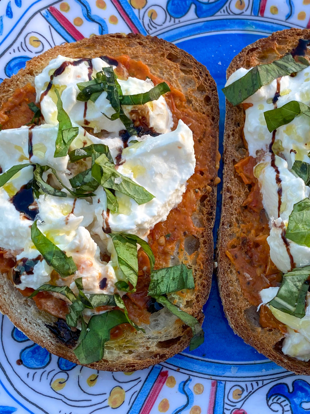 overhead of roasted tomato spread on toast, topped with burrata, drizzled with balsamic vinegar and olive oil, garnished with fresh basil and salt
