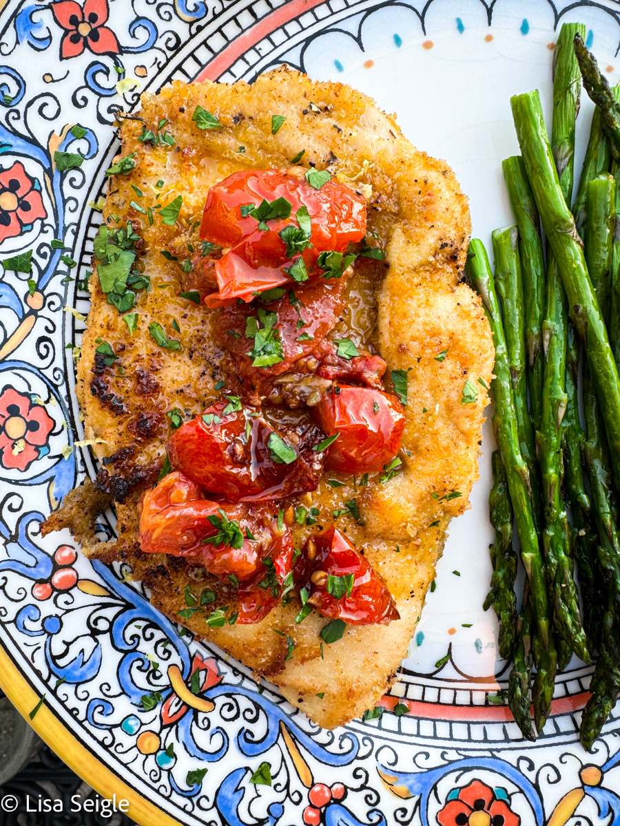 Overhead shot of breaded chicken with asparagus, topped with tomato confit on patterned plate