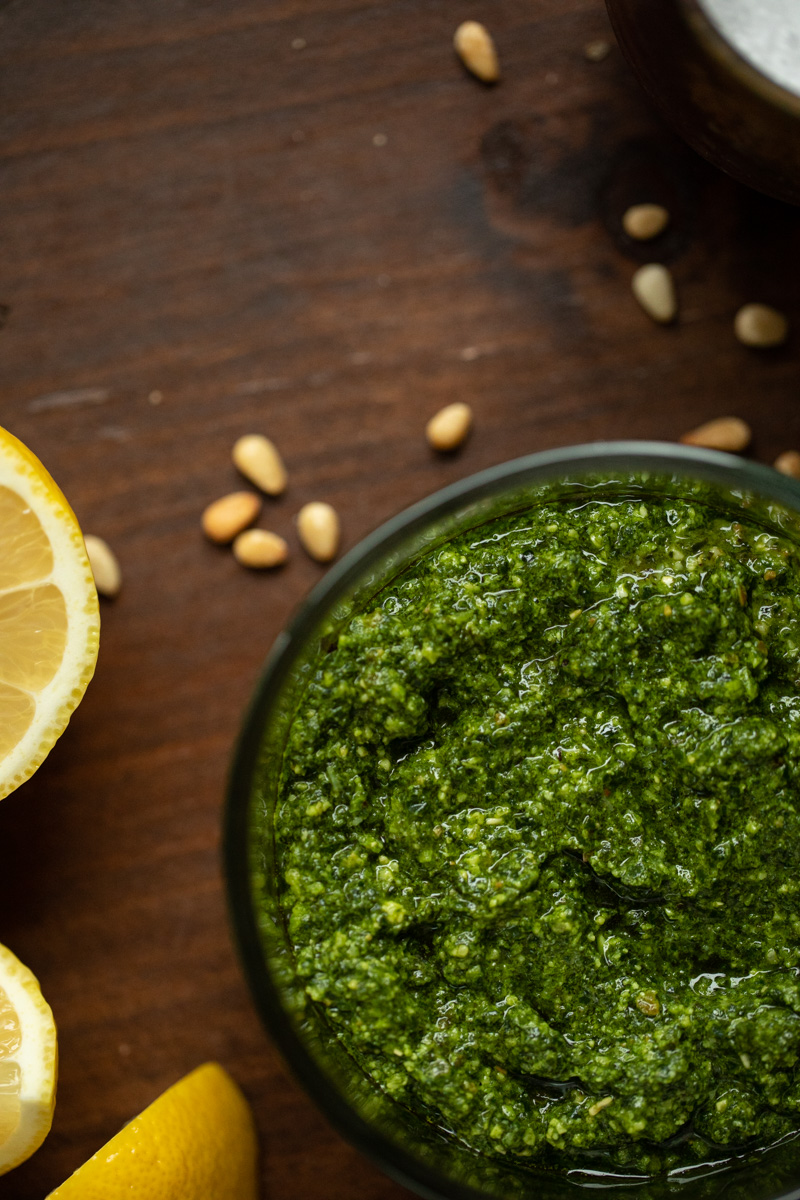 Overhead photo of kale pesto in glass jar on wood board with pine nuts and cut lemon