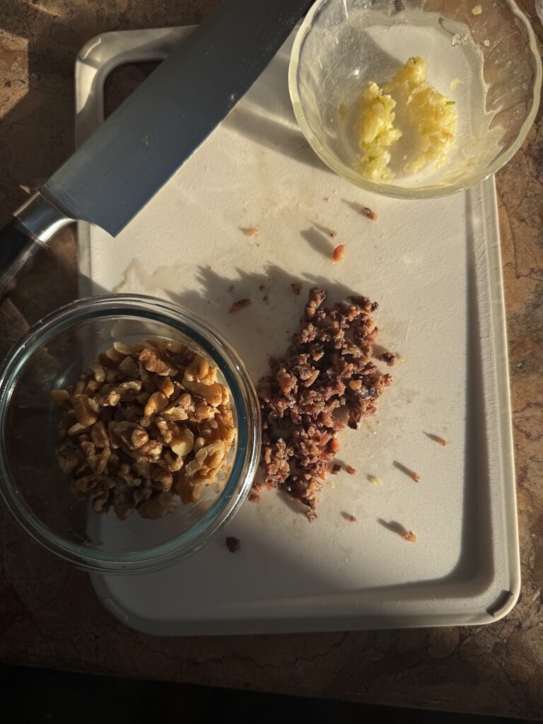minced garlic in small glass bowl, chopped walnuts in small glass bowl and chopped anchovies all on white cutting board with knife.
