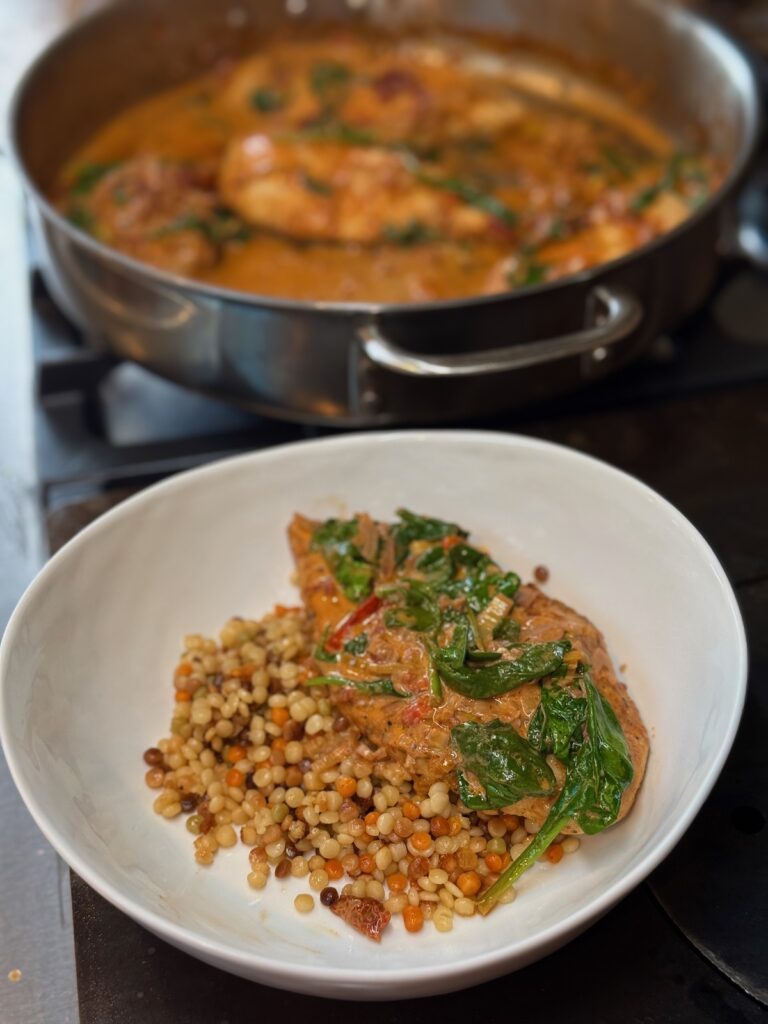 chicken in sundried tomato and spinach sauce in skillet in the background with a serving of chicken in a white bowl over couscous