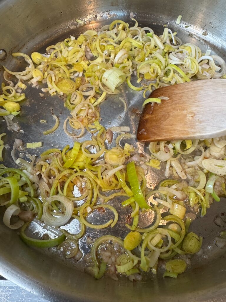 chopped leeks sautéeing in a stainless steel pan and being stirred with a wooden spoon